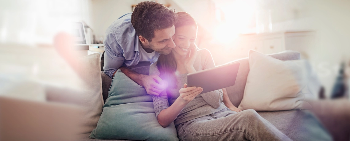 Lächelndes Paar sitzt auf dem Sofa und schaut gemeinsam auf ein Tablet, Sonnenlicht scheint durch das Fenster.