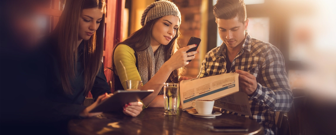 Drei junge Menschen sitzen in einem Café, lesen Zeitung, schauen aufs Tablet und Smartphone, mit Getränken auf dem Tisch.