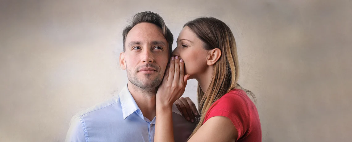 Frau flüstert einem Mann ein Geheimnis ins Ohr, während er nachdenklich schaut.